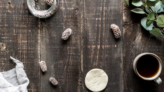 A rustic wooden table features an arrangement of natural elements including small pine cones, a clay disc, and a cup of coffee. A glass jar contains additional pine cones, and a sprig of leaves decorates the right side. A gray cloth napkin is draped to the left.