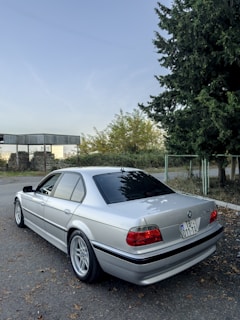 A silver BMW sedan is parked on a quiet street. The background features trees and a small roofed structure. The car's rear is visible, showing a license plate and tinted windows. The area is calm, with fallen leaves on the ground.