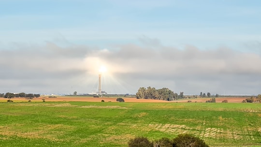 A wide landscape featuring a large field with lush green grass in the foreground. In the distance, there is a solar power tower surrounded by several rows of solar panels. The tower is brightly illuminated at the top, casting a glow that contrasts with the mostly cloudy sky. Several clusters of trees are scattered across the fields.