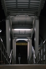 A stairway is illuminated by overhead fluorescent lights, casting a harsh white glow. A person is descending the stairs, partially shadowed, walking on a concrete surface. Metal railings line the sides, and a yellow sign with text is visible in the background, indicating direction.