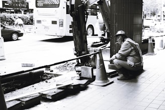 A construction worker wearing a hard hat and uniform is crouching beside an excavator on a tiled sidewalk, taking a break and smoking a cigarette. There are traffic cones and a bus in the background, along with some scattered building materials.