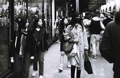 A busy street scene with multiple people walking in both directions. The crowd is diverse, with individuals wearing various winter and fall clothing. Some people are carrying shopping bags, and the large storefront windows display advertisements or photos.