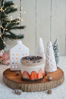 A dessert jar filled with layered cream and strawberry slices sits on a wooden slab. The jar is topped with crushed chocolate or crumb topping. There are small pine cones around the jar, and two decorative white ceramic trees and a miniature white house are placed nearby. A decorated Christmas tree with lights is partially visible in the background.