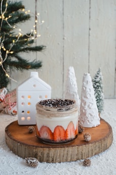 A dessert jar filled with layered cream and strawberry slices sits on a wooden slab. The jar is topped with crushed chocolate or crumb topping. There are small pine cones around the jar, and two decorative white ceramic trees and a miniature white house are placed nearby. A decorated Christmas tree with lights is partially visible in the background.