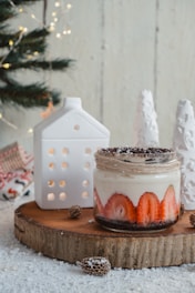 A jar filled with a dessert featuring layers of strawberries and cream is placed on a wooden board dusted with snow-like powder. The setting includes a small, decorative house-shaped candle holder and stylized white trees. A pine branch adorned with fairy lights is visible in the background, enhancing the cozy, holiday ambiance.