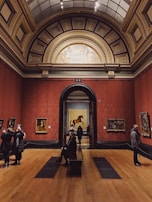 An art gallery with high ornate ceilings and red walls displaying classical artwork. Several people are observing the art pieces, some taking photographs. A large, framed painting of a horse is prominently displayed in the center through an archway.