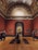 An art gallery with high ornate ceilings and red walls displaying classical artwork. Several people are observing the art pieces, some taking photographs. A large, framed painting of a horse is prominently displayed in the center through an archway.