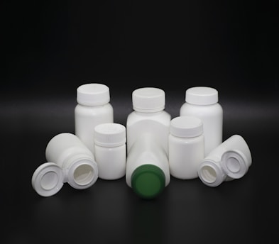 A group of white plastic bottles with various shapes and sizes are arranged on a dark background. Some bottles have screw caps, while others have flip-top lids. The center bottle features a green cap, standing out among the predominantly white lids.