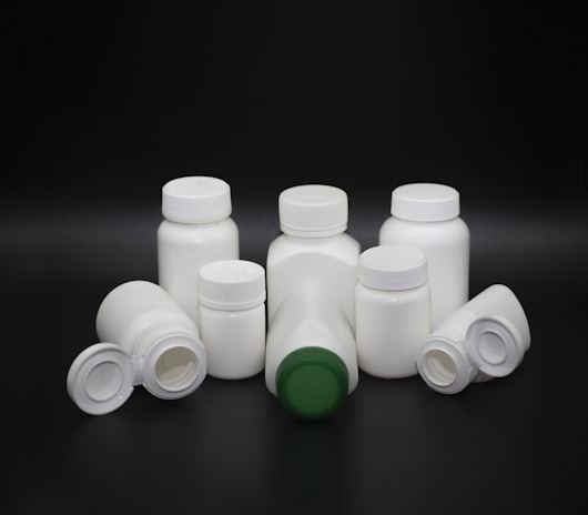 A group of white plastic bottles with various shapes and sizes are arranged on a dark background. Some bottles have screw caps, while others have flip-top lids. The center bottle features a green cap, standing out among the predominantly white lids.