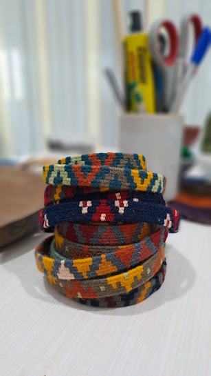 A collection of colorful, woven bracelets with intricate geometric patterns, stacked on a light-colored surface. The background is blurred, showing office or craft supplies such as tape rolls, pens, and a small box.