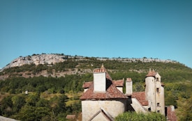 A rustic stone building with red-tiled roofs sits in the foreground. Behind it, a lush green landscape extends to a rocky hillside under a clear blue sky. The building features medieval-style towers and an overall quaint, historical charm.