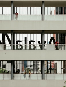 A multi-level structure with people visible on each level. The middle section has large, semi-transparent letters on the windows. The upper and lower sections have individuals standing or walking, with some greenery and urban architecture visible in the background.