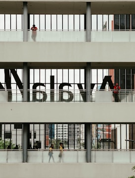 A multi-level structure with people visible on each level. The middle section has large, semi-transparent letters on the windows. The upper and lower sections have individuals standing or walking, with some greenery and urban architecture visible in the background.
