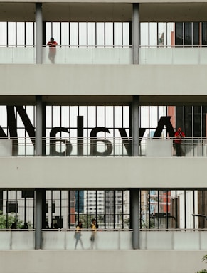 A multi-level structure with people visible on each level. The middle section has large, semi-transparent letters on the windows. The upper and lower sections have individuals standing or walking, with some greenery and urban architecture visible in the background.