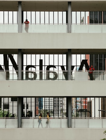 A multi-level structure with people visible on each level. The middle section has large, semi-transparent letters on the windows. The upper and lower sections have individuals standing or walking, with some greenery and urban architecture visible in the background.