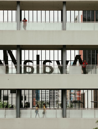 A multi-level structure with people visible on each level. The middle section has large, semi-transparent letters on the windows. The upper and lower sections have individuals standing or walking, with some greenery and urban architecture visible in the background.