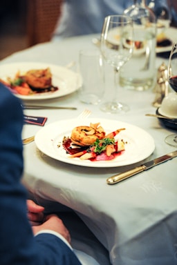 A close-up of a beautifully plated gourmet dish on a white plate, featuring a circular pastry or meat item garnished with fresh herbs and vegetables. The setting appears to be a fine dining restaurant, with elegant table settings including wine glasses, cutlery, and another plate in the background. The tablecloth is light-colored and the overall ambiance suggests a sophisticated dining experience.