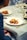 A close-up of a beautifully plated gourmet dish on a white plate, featuring a circular pastry or meat item garnished with fresh herbs and vegetables. The setting appears to be a fine dining restaurant, with elegant table settings including wine glasses, cutlery, and another plate in the background. The tablecloth is light-colored and the overall ambiance suggests a sophisticated dining experience.