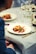A close-up of a beautifully plated gourmet dish on a white plate, featuring a circular pastry or meat item garnished with fresh herbs and vegetables. The setting appears to be a fine dining restaurant, with elegant table settings including wine glasses, cutlery, and another plate in the background. The tablecloth is light-colored and the overall ambiance suggests a sophisticated dining experience.
