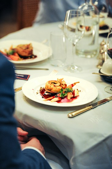 A close-up of a beautifully plated gourmet dish on a white plate, featuring a circular pastry or meat item garnished with fresh herbs and vegetables. The setting appears to be a fine dining restaurant, with elegant table settings including wine glasses, cutlery, and another plate in the background. The tablecloth is light-colored and the overall ambiance suggests a sophisticated dining experience.