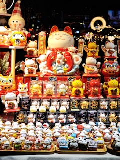 A display of various Maneki-neko figurines, commonly known as lucky cats, arranged in a vibrant market setting. The figurines are of different sizes and colors, predominantly featuring white, red, and gold. Among them is a large, smiling Maneki-neko in the center, surrounded by smaller ones with raised paws. The background showcases a festive and decorative atmosphere with lights and ornaments, hinting at a holiday or cultural celebration.