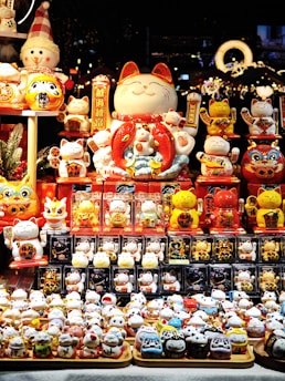 A display of various Maneki-neko figurines, commonly known as lucky cats, arranged in a vibrant market setting. The figurines are of different sizes and colors, predominantly featuring white, red, and gold. Among them is a large, smiling Maneki-neko in the center, surrounded by smaller ones with raised paws. The background showcases a festive and decorative atmosphere with lights and ornaments, hinting at a holiday or cultural celebration.