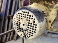 A close-up view of a heavily dust-covered industrial machine part with perforations. The surface appears coarse and textured, with layers of fine dust or sawdust accumulated.