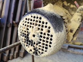 A close-up view of a heavily dust-covered industrial machine part with perforations. The surface appears coarse and textured, with layers of fine dust or sawdust accumulated.