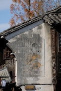 A traditional building with a mural painting on its exterior wall. The mural depicts a person dressed in traditional attire, including detailed accessories like earrings. The building’s architecture features wooden windows and a tiled roof, and the scene is set against a background of trees with autumn foliage.