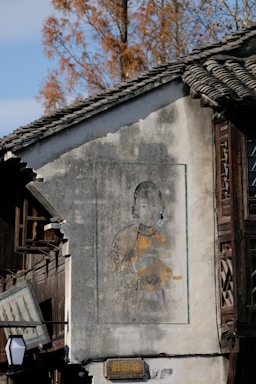 A traditional building with a mural painting on its exterior wall. The mural depicts a person dressed in traditional attire, including detailed accessories like earrings. The building’s architecture features wooden windows and a tiled roof, and the scene is set against a background of trees with autumn foliage.