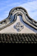 An ornate, traditional architectural roof with intricate designs and patterns. The roof features a carved geometric decoration, and the shingles display a wavy, textured pattern. The sky is clear and blue, creating a contrast with the dark tiles.