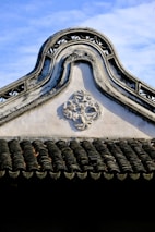 An ornate, traditional architectural roof with intricate designs and patterns. The roof features a carved geometric decoration, and the shingles display a wavy, textured pattern. The sky is clear and blue, creating a contrast with the dark tiles.