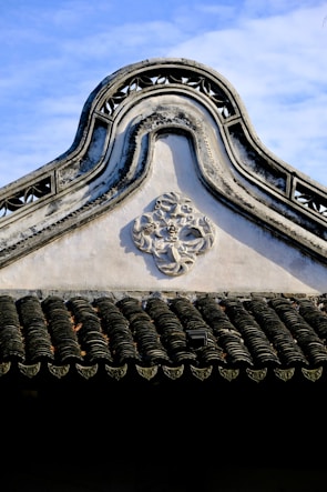 An ornate, traditional architectural roof with intricate designs and patterns. The roof features a carved geometric decoration, and the shingles display a wavy, textured pattern. The sky is clear and blue, creating a contrast with the dark tiles.