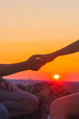 A close-up of two hands reaching through a fence, fingers almost touching, bathed in warm sunset light.