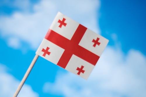 A small flag with a white background and a large red cross is centered in the image. Each quadrant formed by the cross contains a smaller red cross. The flag is held on a stick and is set against a backdrop of a clear blue sky with fluffy white clouds.