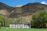 A large country house with a dark roof and symmetrical white-framed windows is set in a green lawn surrounded by deciduous trees. Behind the house towers a rugged, rocky mountain, partially covered with sparse vegetation, under a clear blue sky.