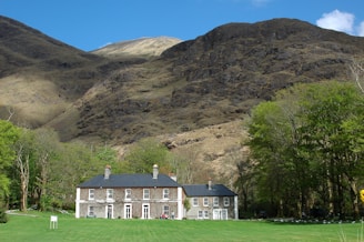 A large country house with a dark roof and symmetrical white-framed windows is set in a green lawn surrounded by deciduous trees. Behind the house towers a rugged, rocky mountain, partially covered with sparse vegetation, under a clear blue sky.