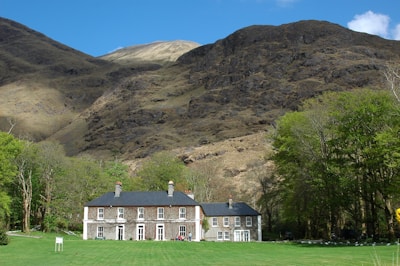 A large country house with a dark roof and symmetrical white-framed windows is set in a green lawn surrounded by deciduous trees. Behind the house towers a rugged, rocky mountain, partially covered with sparse vegetation, under a clear blue sky.