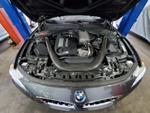 The image captures the engine compartment of a car with an open hood. The engine is prominently displayed, showcasing the intricate details of its components, including various pipes and mechanical parts. The BMW logo is visible on the front of the car, indicating the brand.
