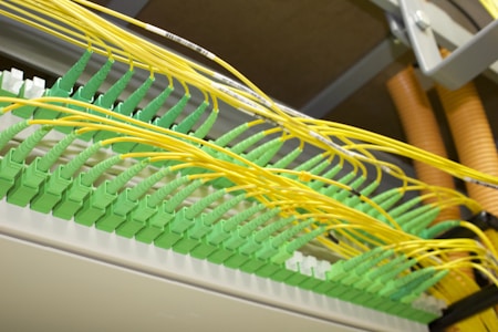A network patch panel with numerous green fiber optic connectors arranged in a row. Bright yellow cables are plugged into each connector, extending outwards in an organized manner. The cables and connectors appear to be part of a structured cabling system in a data center or similar technical environment.