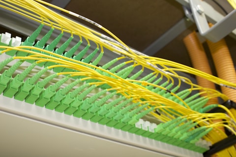 A network patch panel with numerous green fiber optic connectors arranged in a row. Bright yellow cables are plugged into each connector, extending outwards in an organized manner. The cables and connectors appear to be part of a structured cabling system in a data center or similar technical environment.