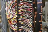 A detailed network or electronic panel features a complex array of multicolored wires organized in rows, connected to a central frame with labeled indicators. The labels on the frame, such as HF and DBRY, suggest its use in telecommunications or electronics. The arrangement of wires indicates intricate circuitry or data connection points.