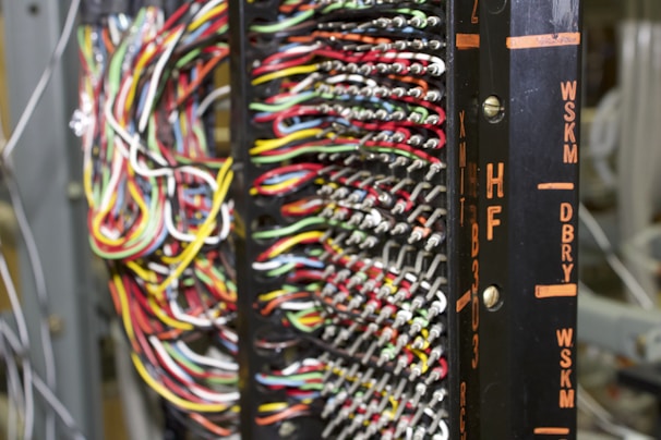 A detailed network or electronic panel features a complex array of multicolored wires organized in rows, connected to a central frame with labeled indicators. The labels on the frame, such as HF and DBRY, suggest its use in telecommunications or electronics. The arrangement of wires indicates intricate circuitry or data connection points.