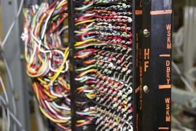 A detailed network or electronic panel features a complex array of multicolored wires organized in rows, connected to a central frame with labeled indicators. The labels on the frame, such as HF and DBRY, suggest its use in telecommunications or electronics. The arrangement of wires indicates intricate circuitry or data connection points.
