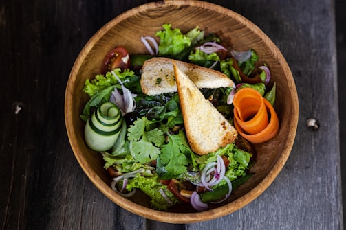 A wooden bowl filled with a vibrant salad. The salad contains fresh leafy greens, thinly sliced red onions, tomato slices, cucumber spirals, and an artistic carrot twirl. Two toasted bread triangles are placed on top, adding a crunchy element.