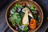 A vibrant salad served in a wooden bowl, featuring a mix of fresh greens, slices of red onion, cherry tomatoes, and a rolled cucumber. Two pieces of toasted bread are placed on top, with a garnish of curled carrot slices.