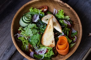 A vibrant salad served in a wooden bowl, featuring a mix of fresh greens, slices of red onion, cherry tomatoes, and a rolled cucumber. Two pieces of toasted bread are placed on top, with a garnish of curled carrot slices.