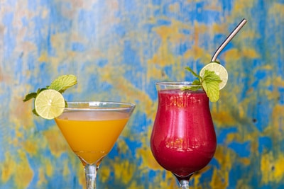 Two colorful cocktails are displayed against a vibrant blue and yellow textured background. The drink on the left is a yellow-orange color, garnished with a lime slice and mint leaves, served in a martini glass. The drink on the right is a deep red-pink color, also garnished with a lime slice and mint, served in a tall glass with a metallic straw.