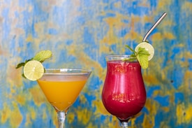 Two colorful cocktails are displayed against a vibrant blue and yellow textured background. The drink on the left is a yellow-orange color, garnished with a lime slice and mint leaves, served in a martini glass. The drink on the right is a deep red-pink color, also garnished with a lime slice and mint, served in a tall glass with a metallic straw.