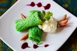 A neatly plated dessert featuring two vibrant green rolled crepes next to a scoop of creamy vanilla ice cream. Fresh mint leaves are placed on top for garnish, while sliced strawberries and drizzles of red sauce add a pop of color on a white square plate.